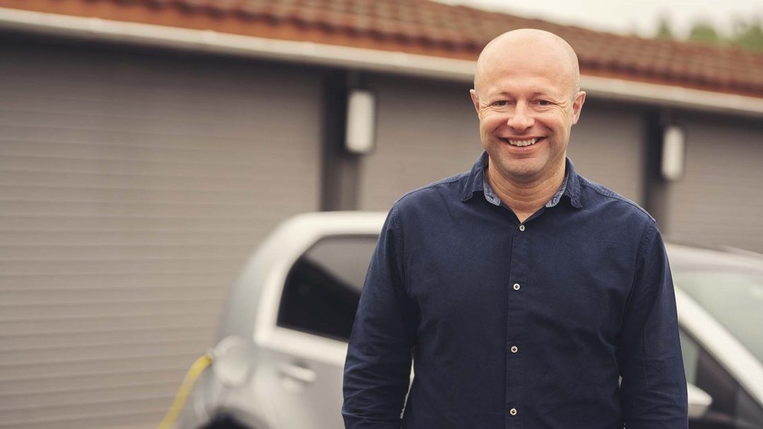 En person i mørk skjorte smiler foran en ladede elbil og garasjeporter.