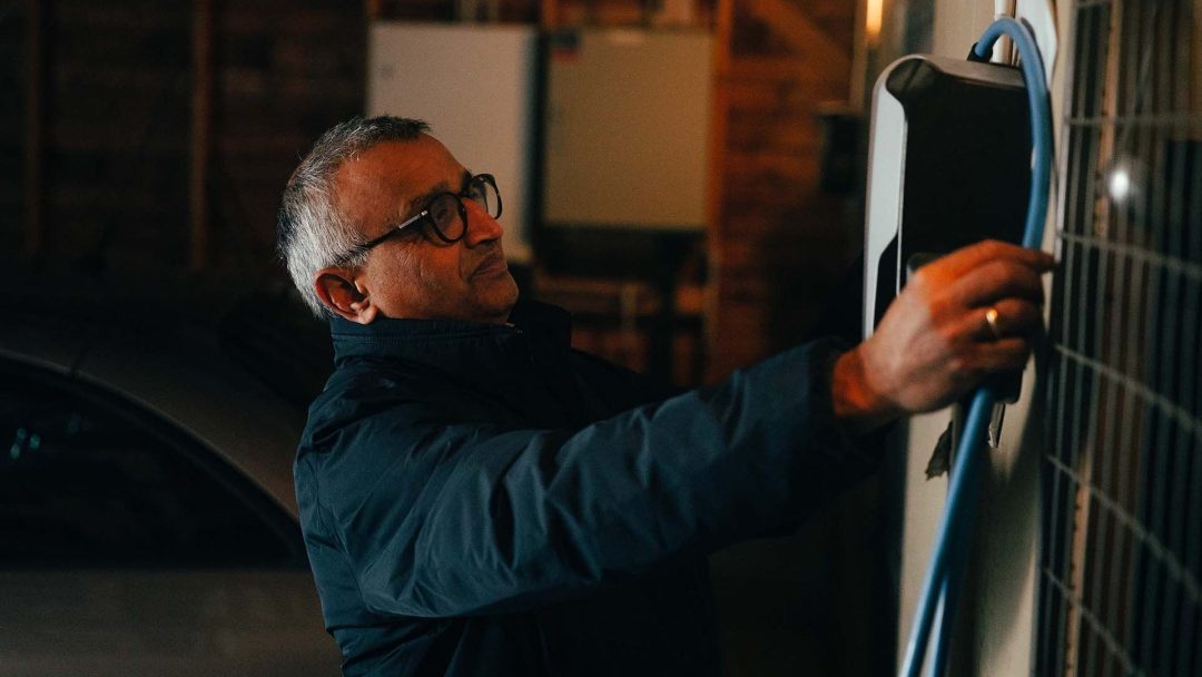 En person med briller og mørk jakke kobler en blå ladekabel til en veggmontert lader i en svakt opplyst garasje.