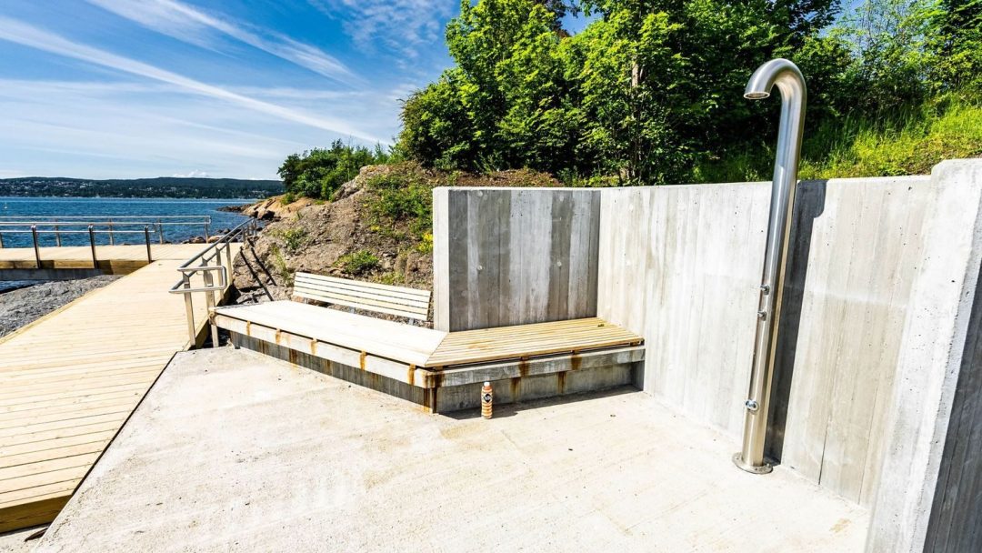 Utendørsdusj med metallarmatur nær trebenker på betongplattform, som minner om sjarmerende badeplasser. Det er omgitt av frodige grøntområder, og tilbyr en rolig utsikt over havet og fjerne åser under en pulserende blå himmel.