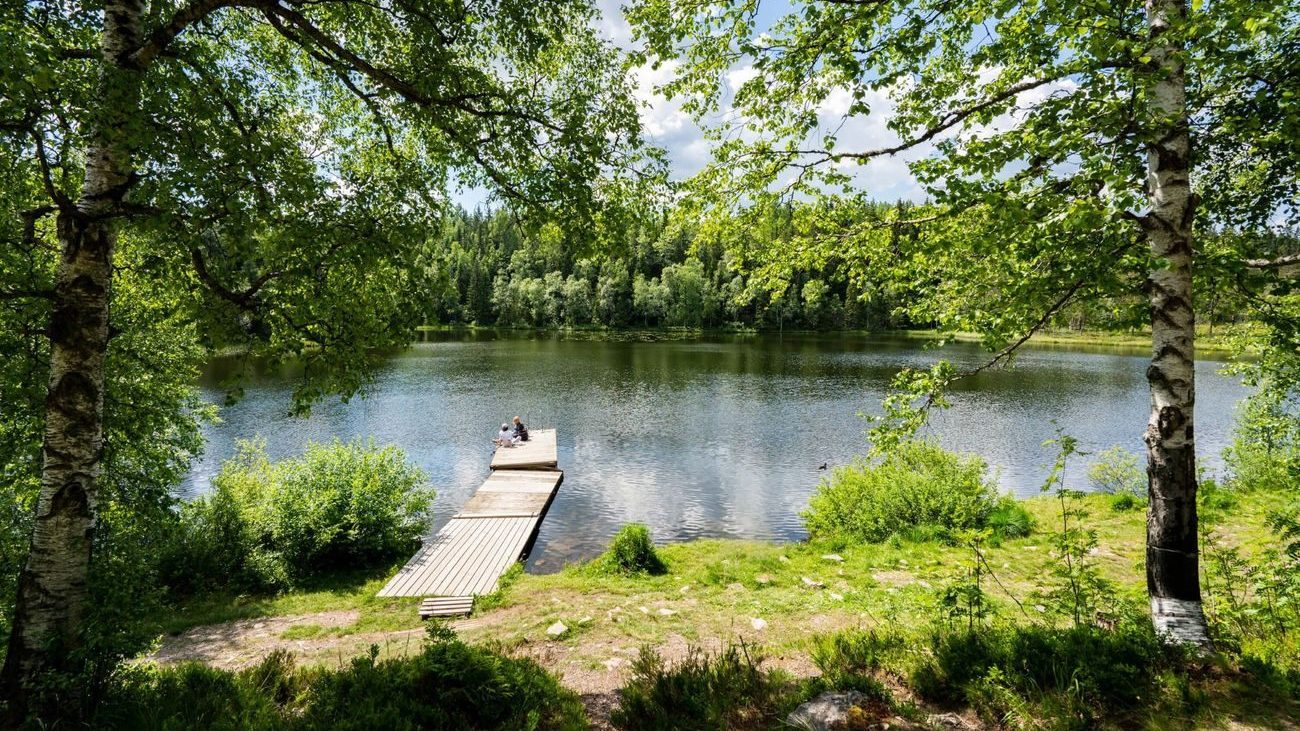 To personer sitter på en trebrygge som strekker seg ut i en rolig innsjø, omfavnet av frodige grønne trær under en delvis overskyet himmel – et perfekt badeplasser, hvor naturens ro inviterer til fredelig ettertanke.