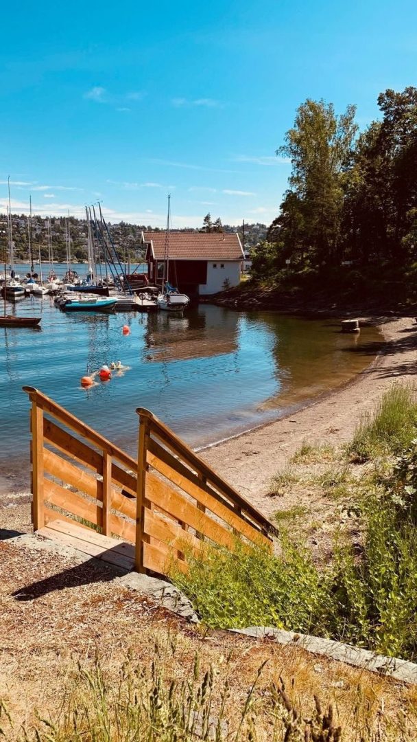 Tretrapper fører ned til sandstrande badeplasser ved en rolig bukt med fortøyde seilbåter og et naust med rødt tak, omgitt av trær under en klarblå himmel.