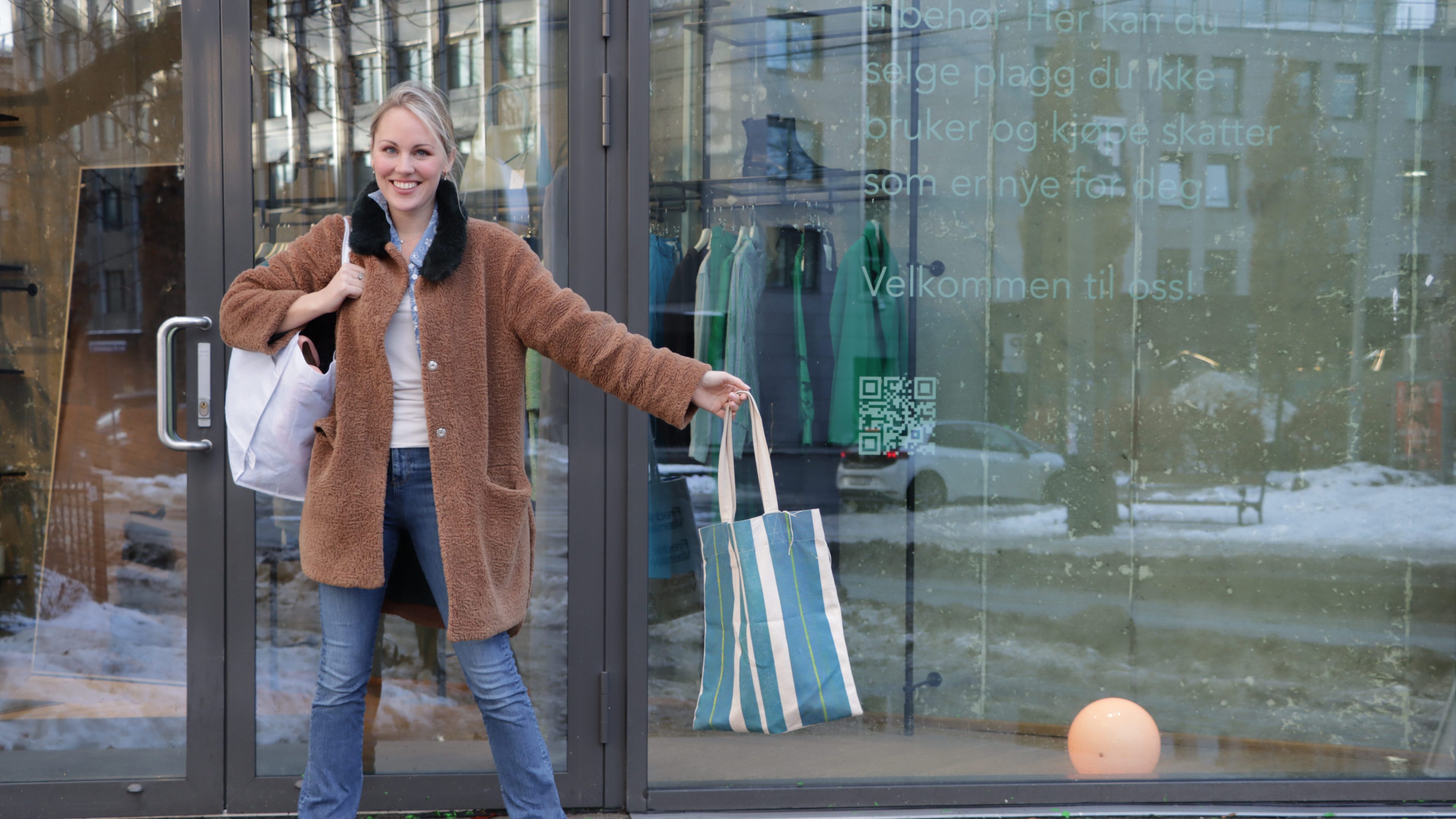 En person i brun frakk står utenfor en glassbutikk på bruktbutikker, holder to poser og smiler. Snø dekker bakken og bidrar til den koselige atmosfæren.