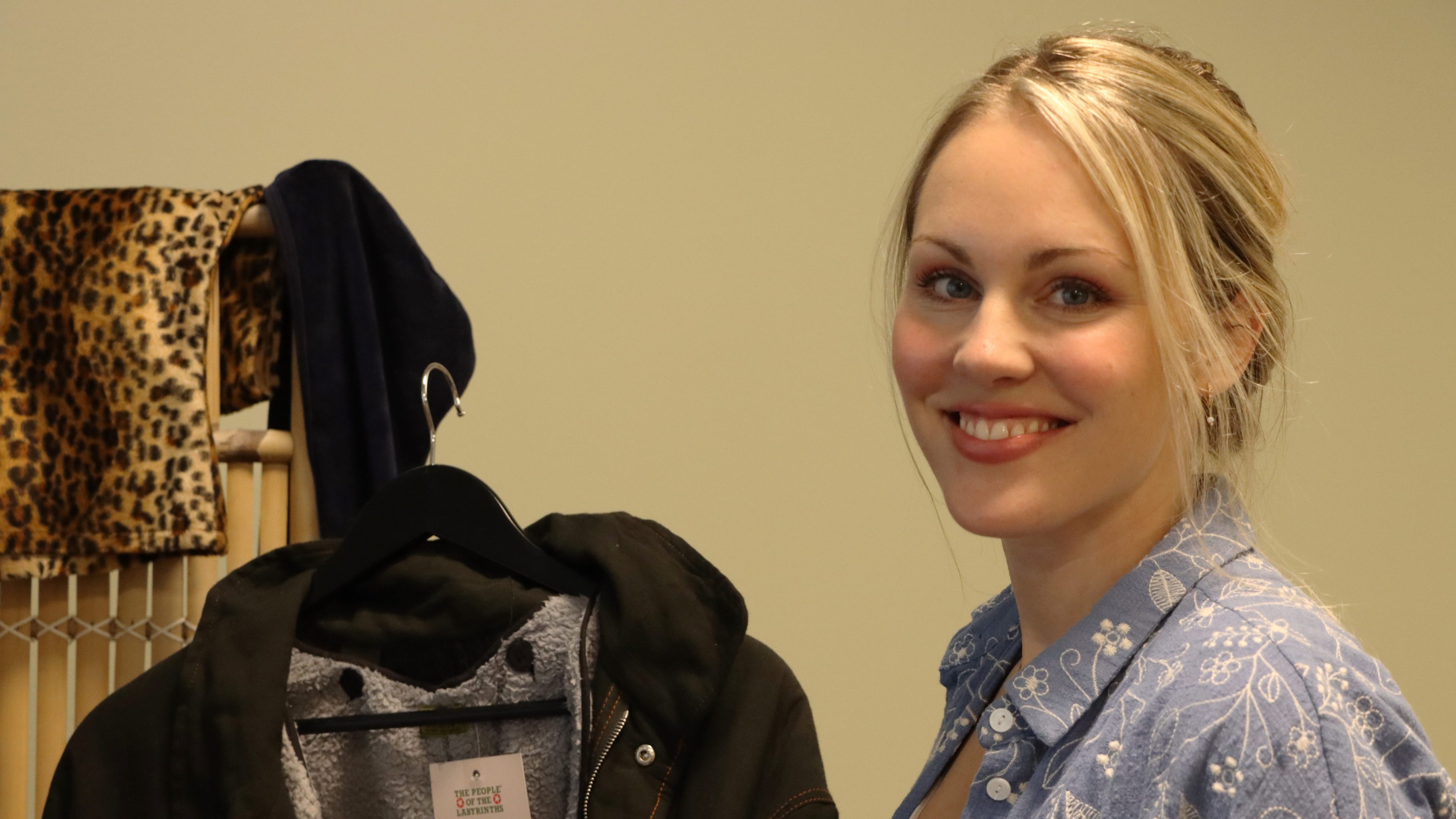 En smilende person står innendørs iført en blåmønstret skjorte fra en lokal bruktbutikk. I nærheten henger en svart jakke på en stol med et leopardmønster drapert over.