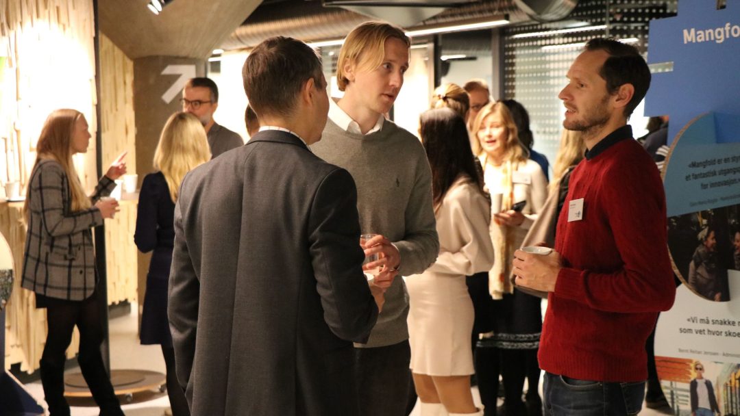 Folk samlet seg innendørs, minglet og holdt drinker. Tre menn samtaler i forgrunnen. Andre er engasjert i samtale i bakgrunnen. Innstillingen ser ut til å være et profesjonelt arrangement eller nettverksøkt.