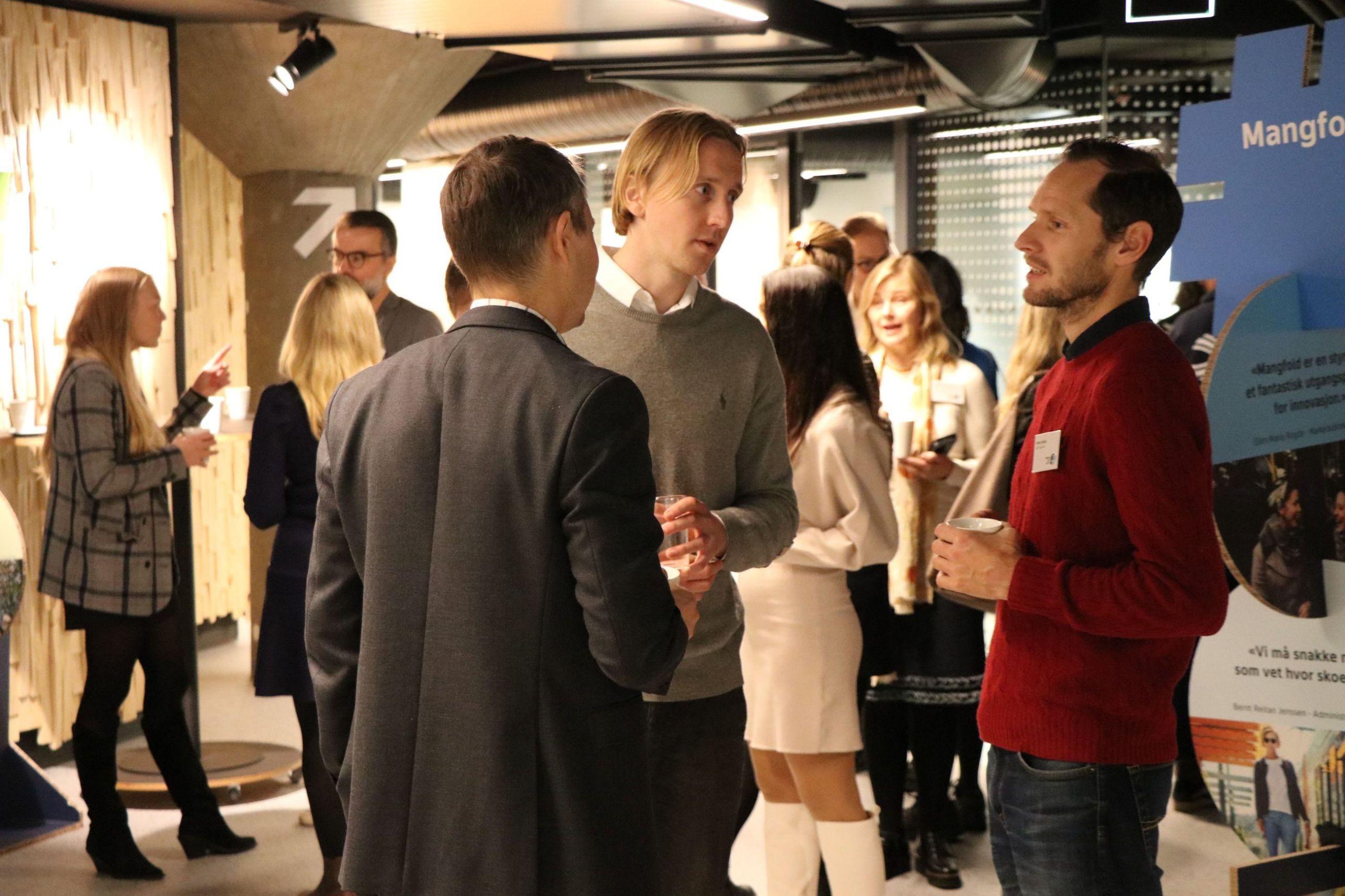 Folk på et nettverksarrangement snakker i grupper og holder drinker. En mann i en grå genser og en annen i en rød genser snakker med en mann i dress. Plakater og en trevegg er synlig.
