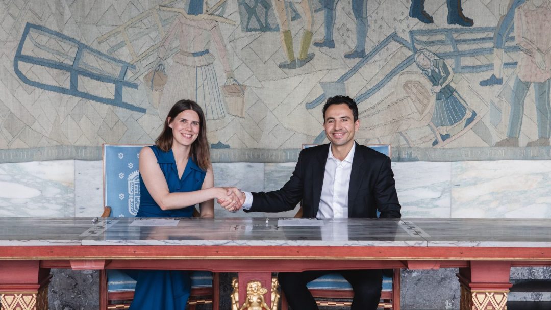 To personer som sitter ved et bord og håndhilser, med et veggmaleri som viser arbeidere i bakgrunnen.