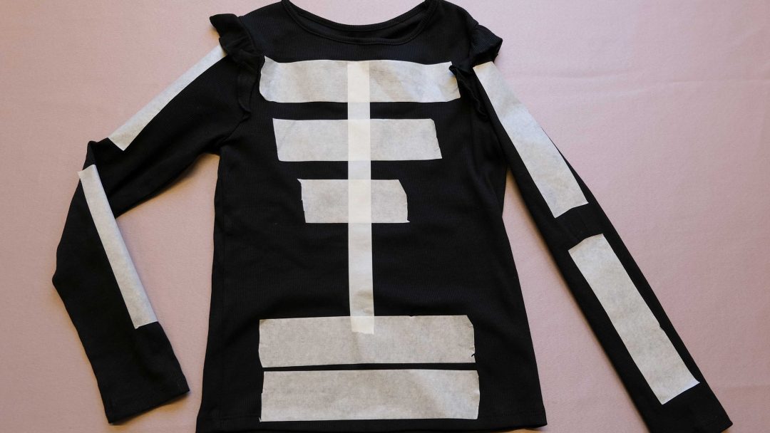 Denne svarte langermede skjorten har hvite papirstrimler kunstferdig arrangert i et skjelettmønster foran og på armene, perfekt for en smart halloweenkostyme. Lagt flatt på en rosa overflate, utstråler den skummel stil med enkelhet.