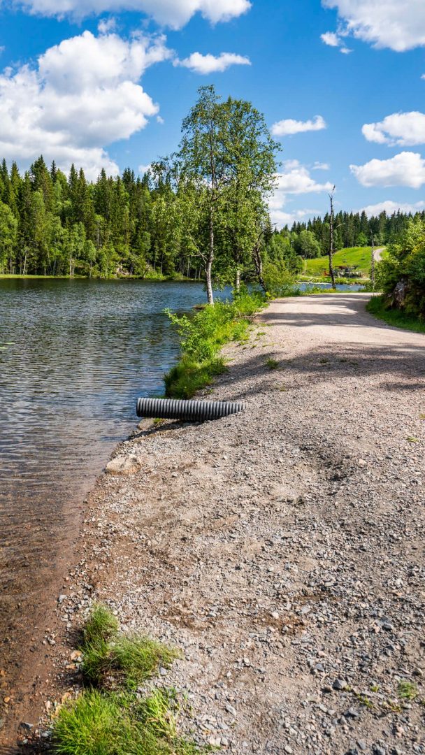 Den rolige stien langs innsjøen, flankert av trær under overskyet himmel, fører til rolige badeplasser. Her hviler et kulvertrør subtilt nær vannkanten.