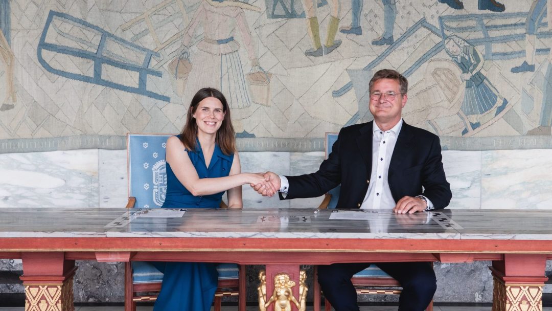 En kvinne og en mann sitter ved et bord, håndhilser og smiler, med dokumenter foran seg. Et dekorativt veggmaleri er i bakgrunnen.