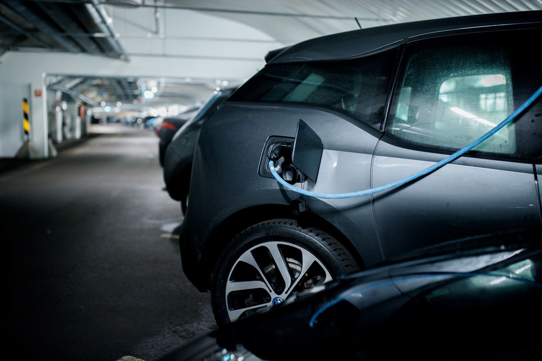 I et svakt opplyst parkeringshus beregnet for «lading av elbil i borettslag» lader en elbil flittig med den blå kabelen sikkert koblet til ladeporten, mens andre kjøretøy står parkert i nærheten.