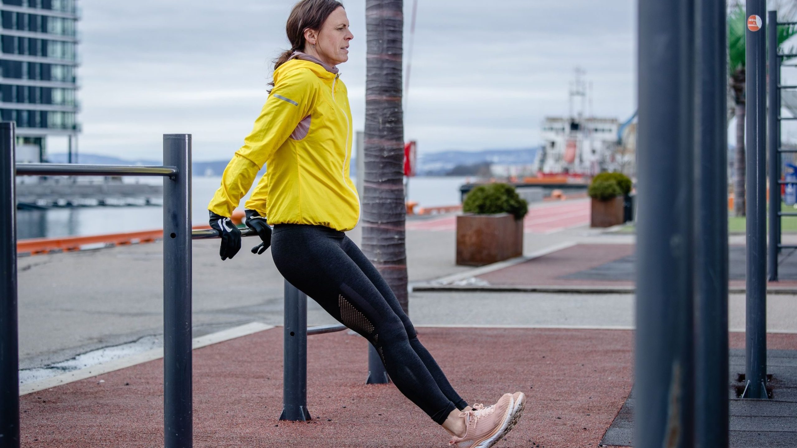 Person i gul jakke og sorte leggings utfører styrketrening med tricep-dips på parallelle stenger utendørs nær en vannkant.