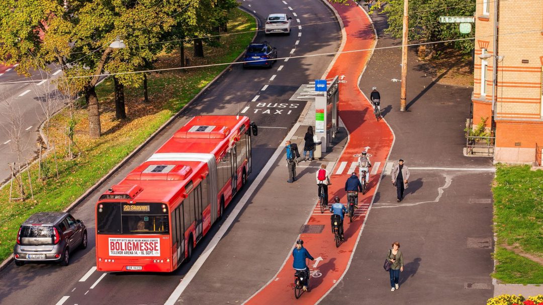 En rød buss venter ved bygatens bussholdeplass, omgitt av sykkelfelt hvor syklister følger sykkelregler. Fotgjengere rusler under trærne, og parkerte biler langs kanten av veien.