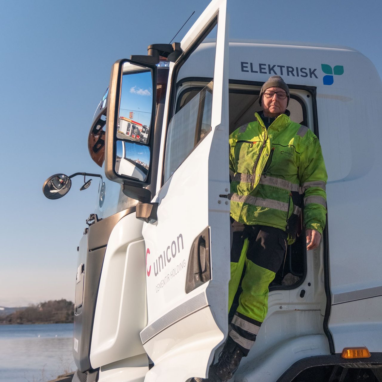 En person i synlige klær står i den åpne døren til en hvit elektrisk betongbil, parkert nær en rolig vannmasse.