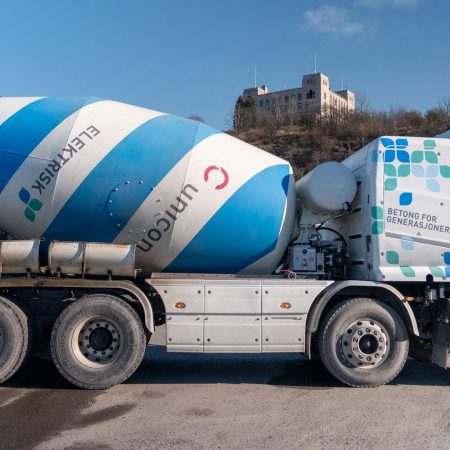 En blå og hvit elektrisk betongbil står parkert på en vei, innrammet av en åsside og bygning i bakgrunnen.