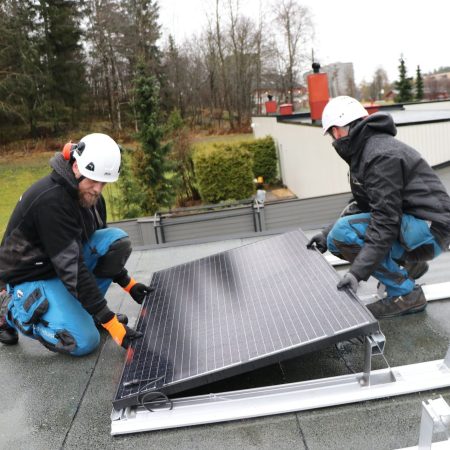 To arbeidere i verneutstyr installerer et solcellepanel på taket, og drar fordel av klimatilskudd-insentiver, omgitt av trær og bygninger i bakgrunnen.