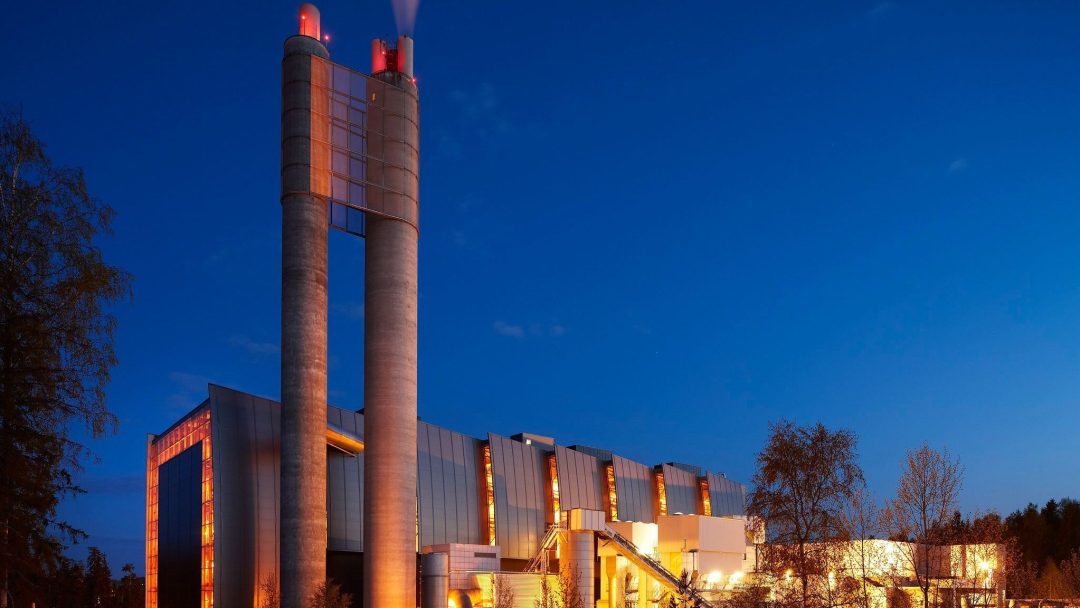 Et moderne industrianlegg med to høye skorsteiner sender ut lys mot en skumringshimmel, omgitt av trær, der selv indirekte utslipp spiller en rolle i kveldsstemningen.