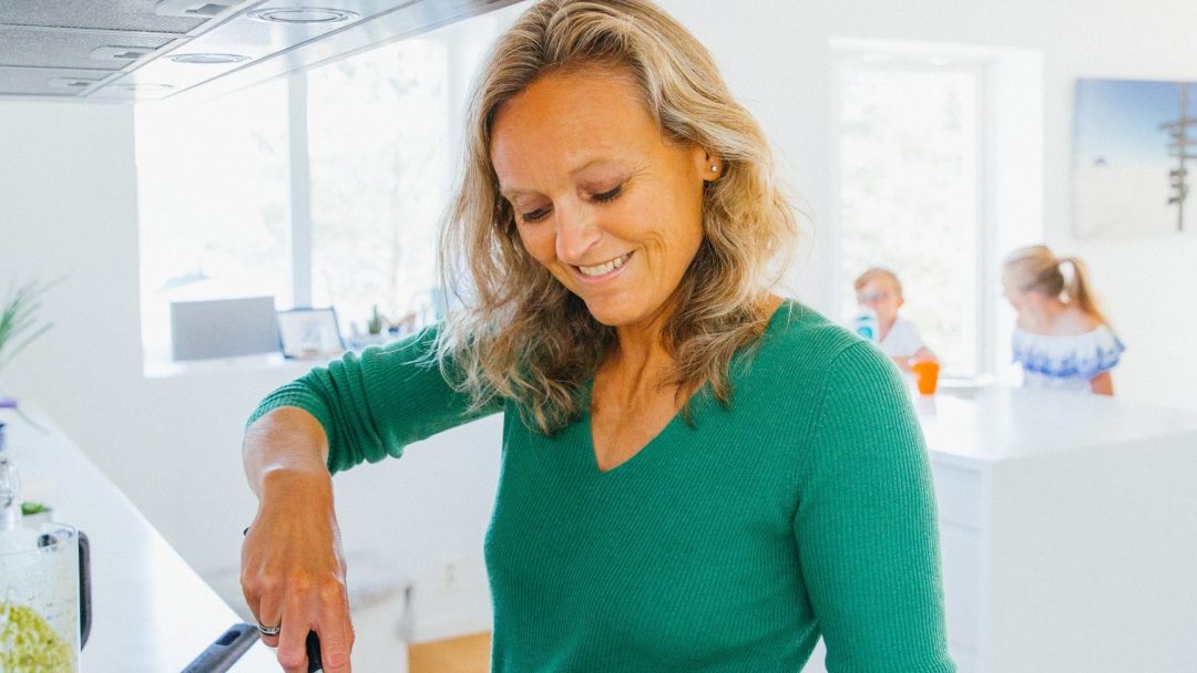 En kvinne i en grønn genser lager mat på et lyst kjøkken og sorterer dyktig avfall, med to personer som sitter ved et bord i bakgrunnen.