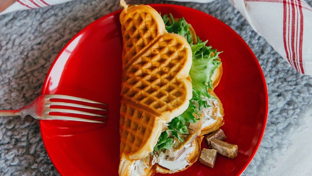 En hjerteformet vaffelsandwich med salat, kjøtt og et snev av avfall på en rød tallerken, akkompagnert av en gaffel på en strukturert overflate.
