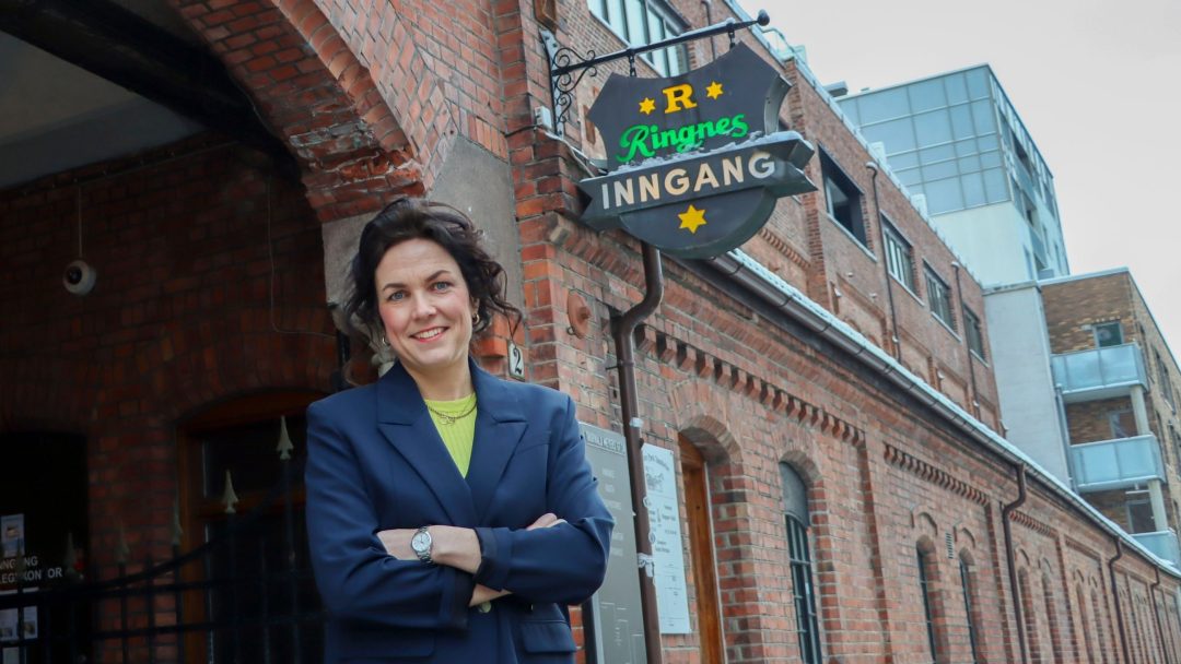 En person i en blå blazer, laget av resirkulert plast, står med armene i kors utenfor en murbygning med et skilt der det står «Ringnes Inngang.