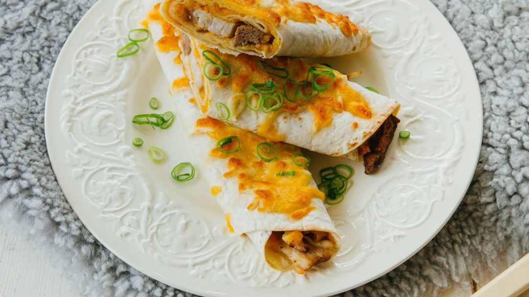 To burritos fylt med kjøtt og toppet med smeltet ost, grønn løk i skiver og et dryss avfall på en dekorativ hvit tallerken.