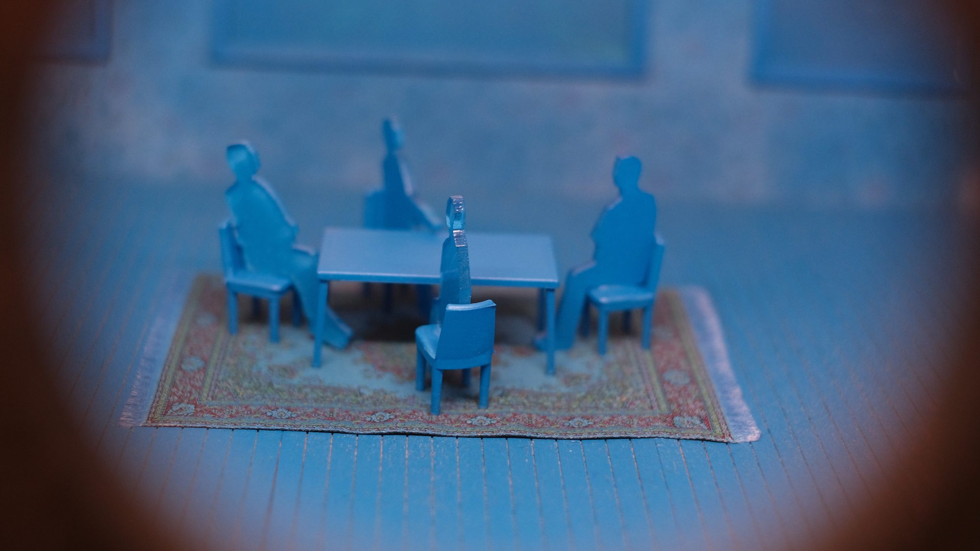 Miniatyrblå figurer som sitter rundt et bord på et mønstret teppe, sett gjennom en sirkulær ramme, gjenspeiler den rolige atmosfæren i Klimahusets rolige utstillinger.