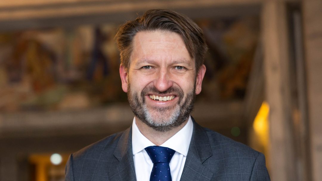 Smilende mann i dress med blått slips, som står innendørs og utstråler New Yorks polerte sjarm.