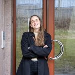 En person med langt hår, iført en svart frakk, står selvsikkert med armene i kors foran en glassdør, og legemliggjør oppførselen til en energirådgiver. Frodig gress og trær skaper et rolig bakteppe.