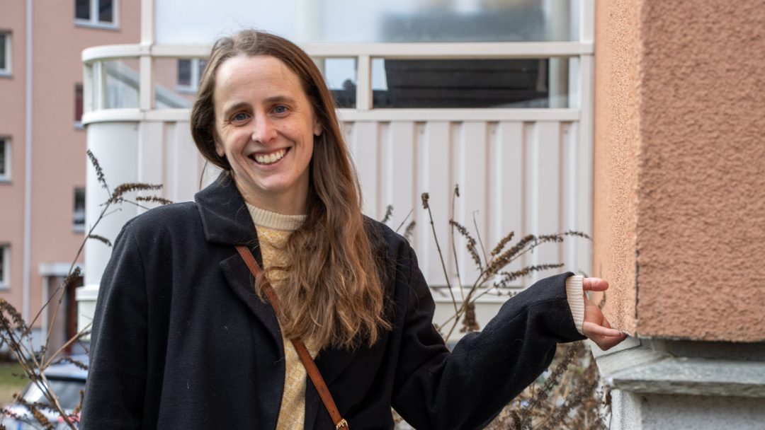 En person med langt hår i svart frakk, som kanskje er en energirådgiver, smiler og peker på en bygning eksteriør med tørkede planter i forgrunnen.