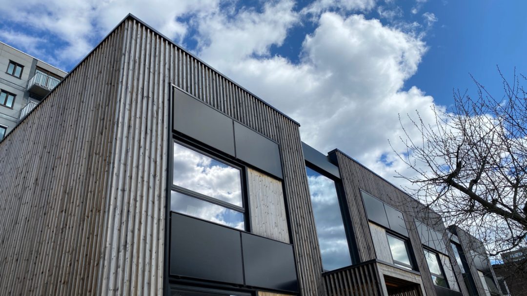 Moderne trebygning med svarte aksenter under en overskyet blå himmel. Bare tregrener på høyre side av bildet.