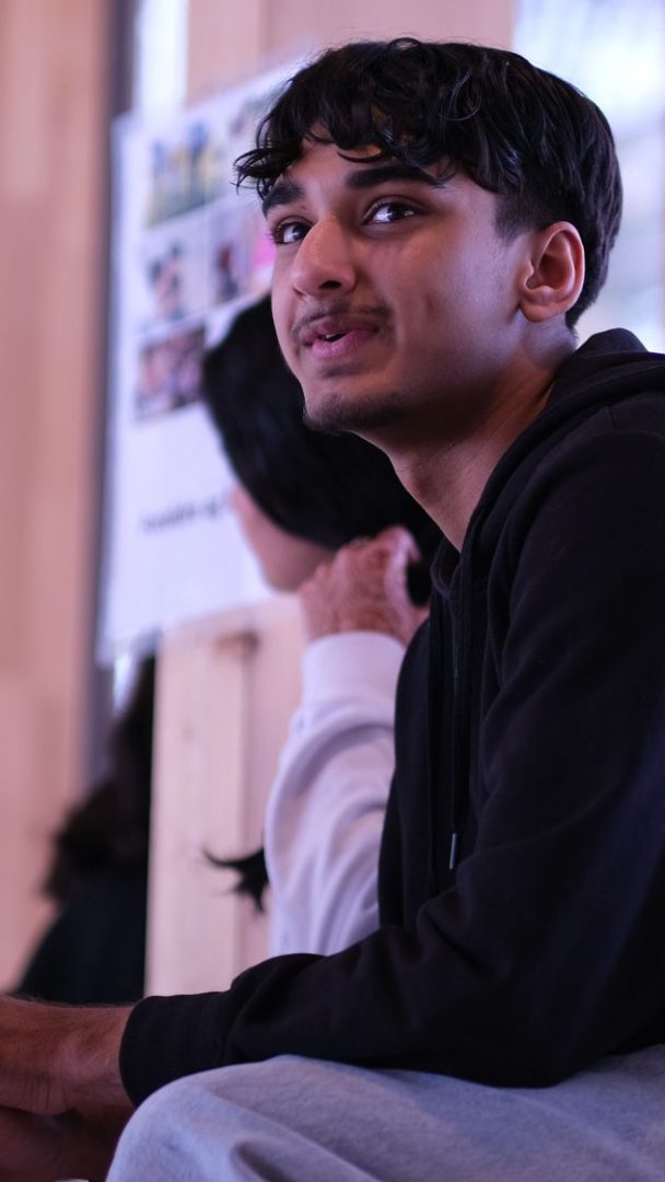 En person med kort, mørkt hår og svart hettegenser sitter innendørs på Klimahuset og ser oppover. Den uskarpe bakgrunnen avslører en annen figur og en plakat.