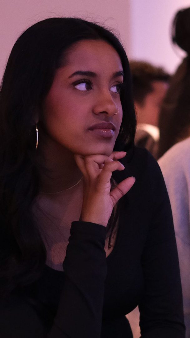 En kvinne med langt mørkt hår, iført en svart topp, hviler haken på hånden mens hun stirrer sidelengs, tilsynelatende fortapt i tankene midt i Klimahusets moderne bakteppe.