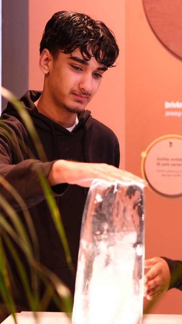 I den rødbrune gløden fra Klimahusets innendørssetting tar en person kledd i en svart hettegenser forsiktig på en stor isblokk som hviler på bordet.