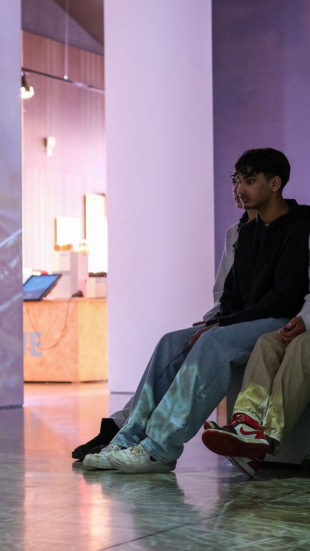 To personer sitter på en benk i Klimahuset, omgitt av moderne arkitektur, med hypnotiserende lysmønstre projisert på gulvet og bena.