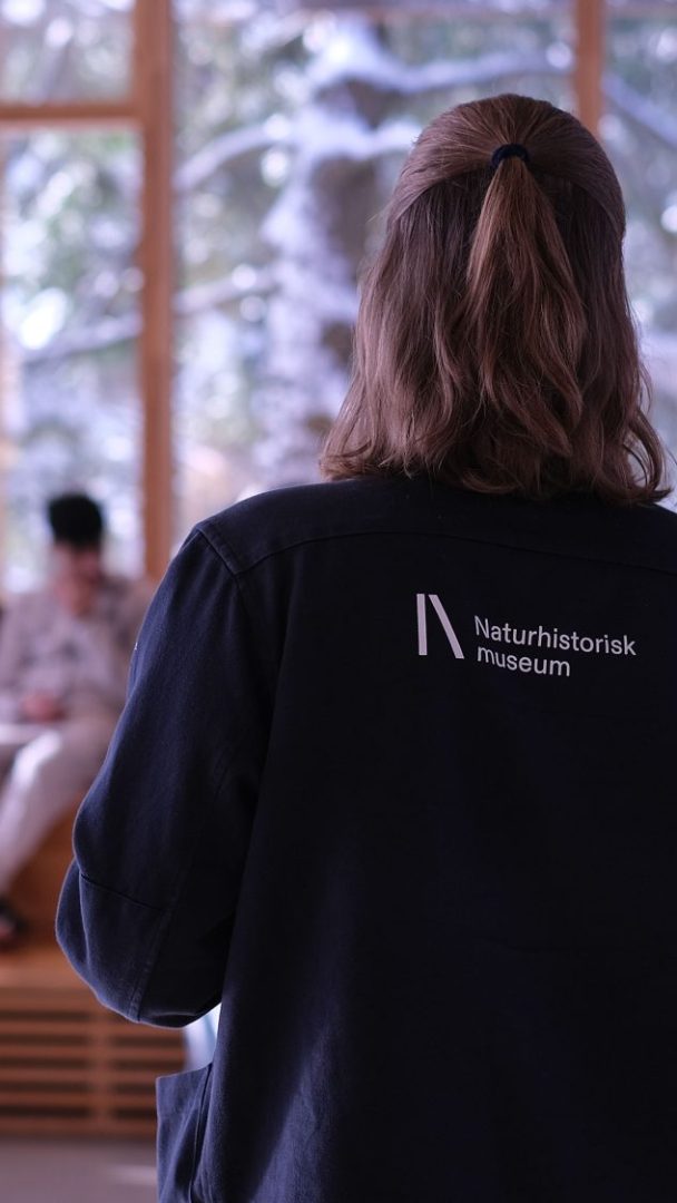 En person er vendt bort, iført jakke med «Naturhistorisk museum»-tekst, inne i Klimahuset. Scenen inkluderer en uskarp figur og snødekte trær synlig gjennom vinduene.