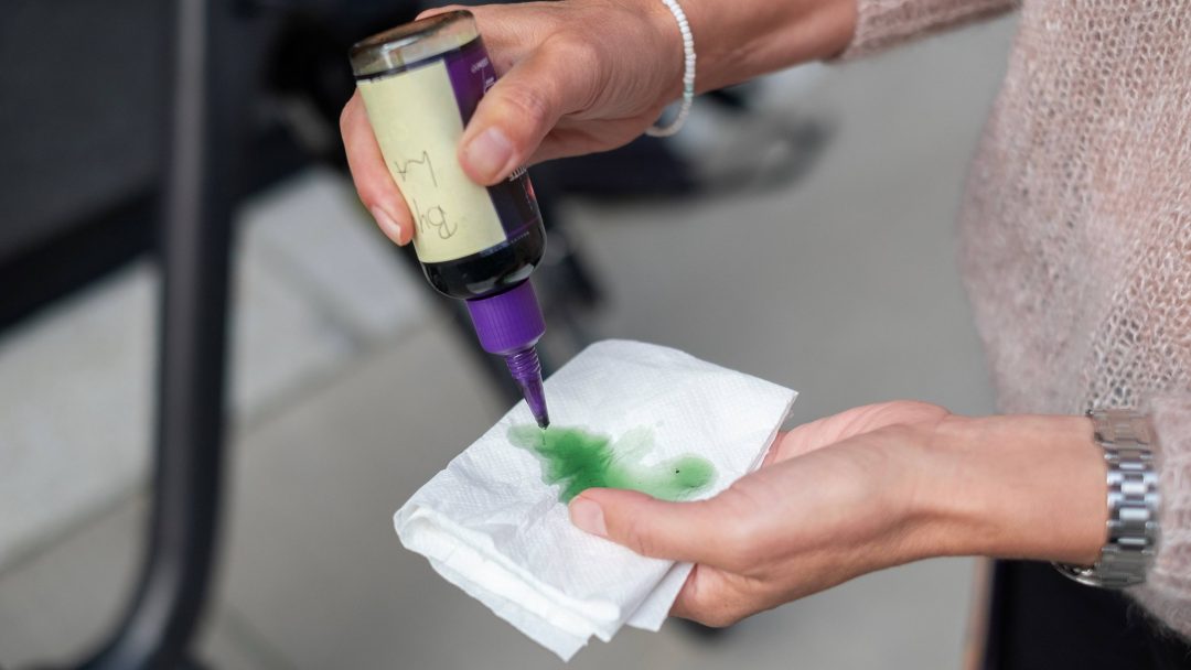 En person heller forsiktig grønn væske, som minner om en fin sykkelvask-løsning, fra en flaske på et hvitt papirhåndkle med begge hender.