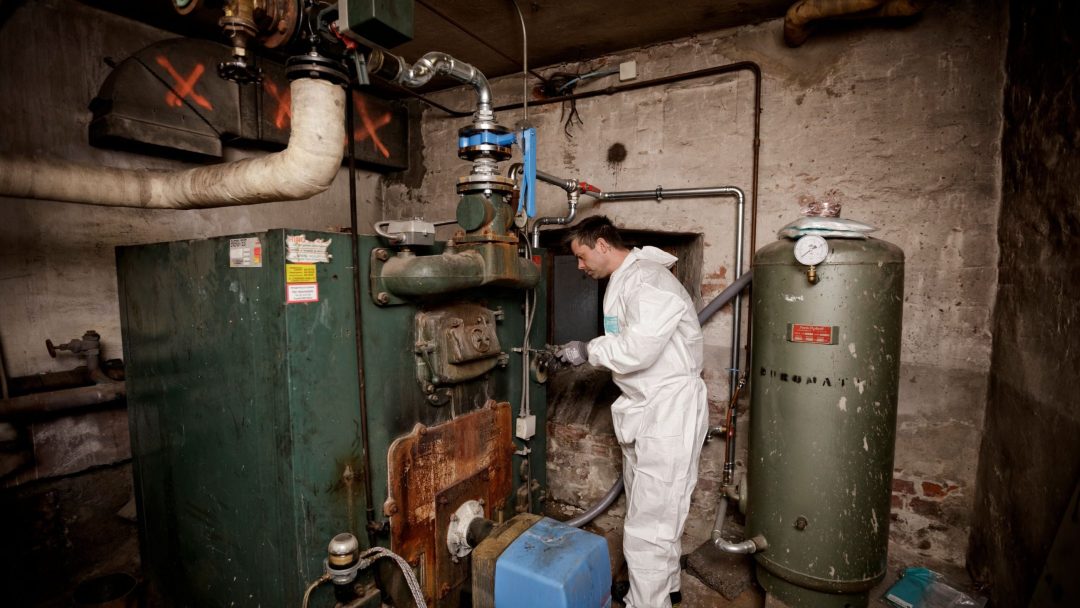 En person i hvit dress undersøker gamle industrimaskineri i et svakt opplyst rom, omgitt av rør og utslippsdekket rustent utstyr.