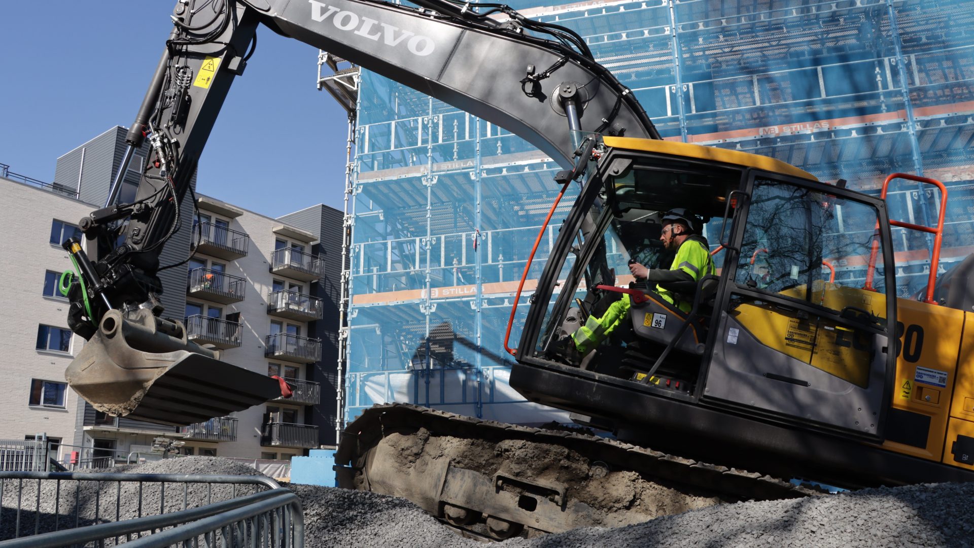 En bygningsarbeider driver en Volvo gravemaskin på en grusplass ved siden av en blå bygning, med moderne leiligheter synlig i bakgrunnen, med tanke på å minimere klimagassutslipp.