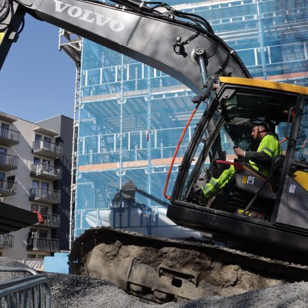 En bygningsarbeider driver en Volvo gravemaskin på en grusplass ved siden av en blå bygning, med moderne leiligheter synlig i bakgrunnen, med tanke på å minimere klimagassutslipp.