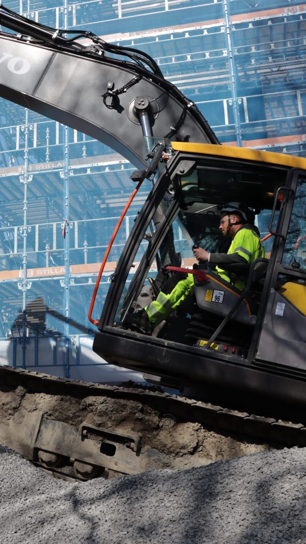 En person betjener en gravemaskin på en byggeplass, og håndterer klimagassutslipp nøye. Maskinen er plassert på grus, med en delvis konstruert bygning dekket av stillas i bakgrunnen.