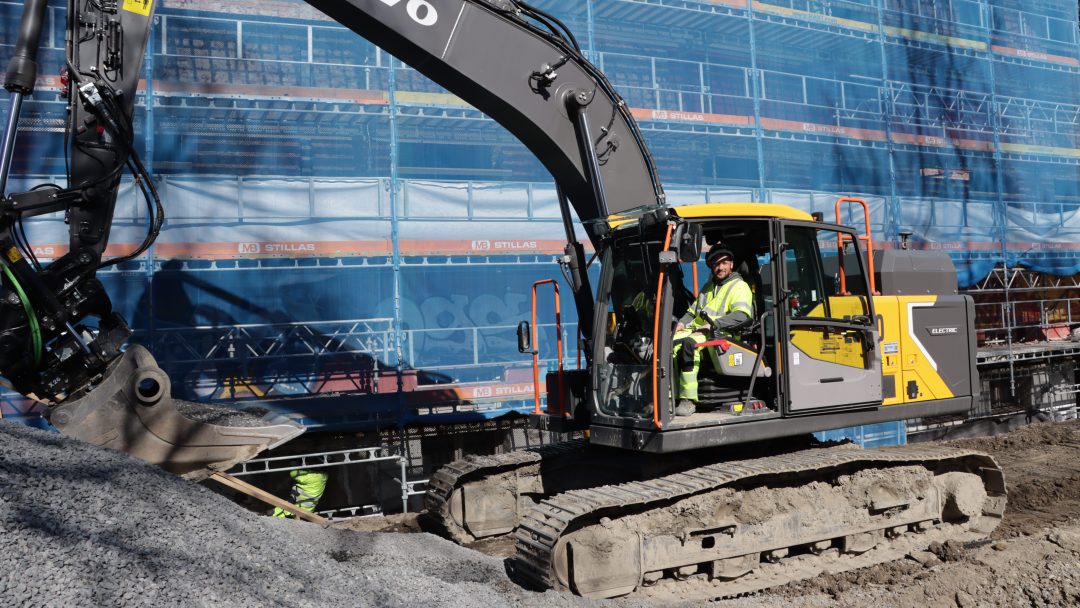 En bygningsarbeider som driver en gravemaskin på en grusplass med en bygning under blå stillaser i bakgrunnen, med tanke på klimagassutslipp mens de jobber med å redusere miljøpåvirkningen.