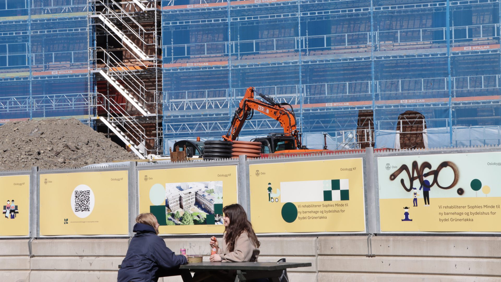 To personer sitter ved et piknikbord foran en byggeplass med kran og stillas, mens skilt på gjerdet fremhever arbeidet med å redusere klimagassutslipp.