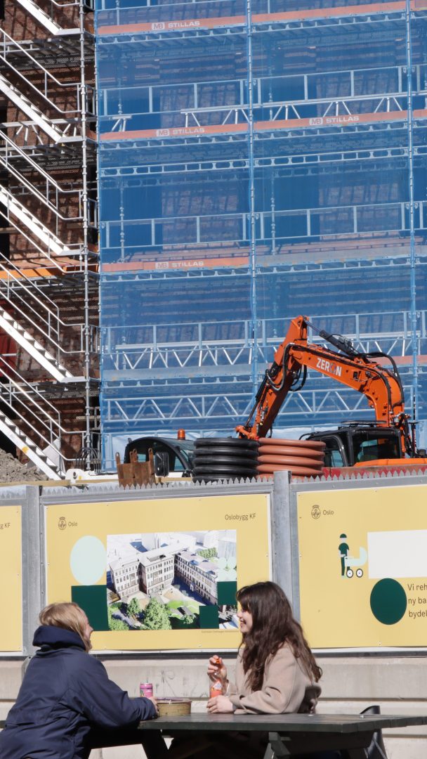 To personer sitter ved et bord foran en byggeplass med stillas og en oransje gravemaskin og diskuterer klimagassutslipp. Et skilt med byggeplaner vises på et gjerde, som fremhever bærekraftig praksis for å redusere utslipp.