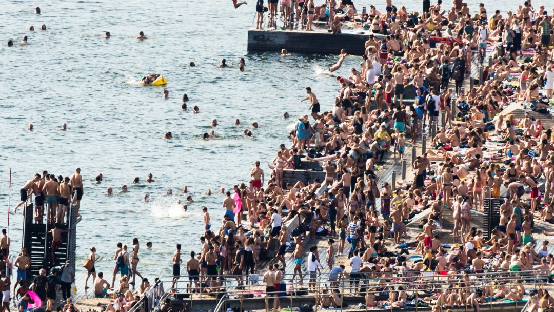 Overfylt strandscene med mange mennesker som svømmer, soler seg og hopper i vannet fra en brygge. Området er spekket med enkeltpersoner som nyter en solrik dag ved sjøen.