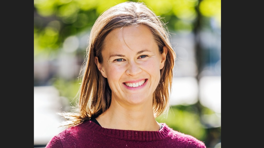 En person med skulderlangt hår smiler mens han står utendørs i en rødbrun genser, omgitt av uskarpt grønt, med tanke på utslippets påvirkning på miljøet.