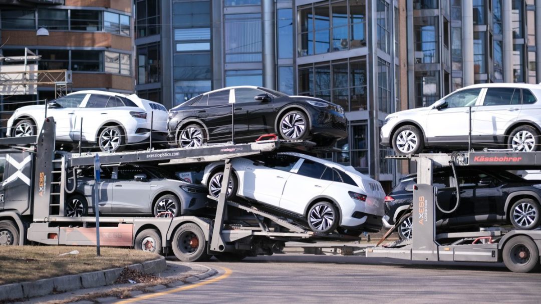 En personbil lastet med flere nye biler står parkert på en gate i nærheten av moderne glassbygninger.