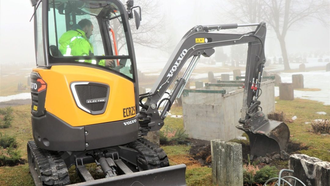 På den tåkete kirkegården betjener en person en gul og svart minigraver, som manøvrerer stille blant gravsteiner og snøflekker, og viser presisjonen samtidig som utslippene forblir minimale.