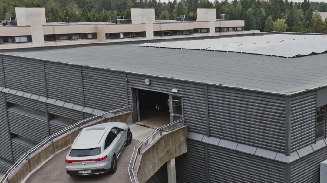 En hvit bil kjører inn i et parkeringshus med flere nivåer omgitt av trær.