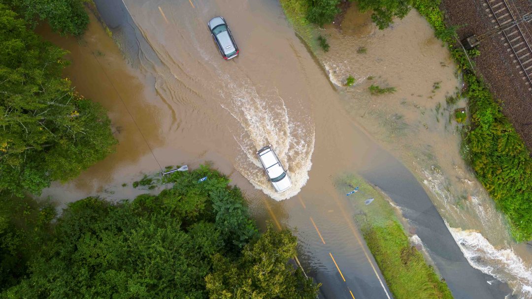 Luftfoto av to biler som kjører gjennom en oversvømmet kjørebane omgitt av grøntområder og et jernbanespor i nærheten.