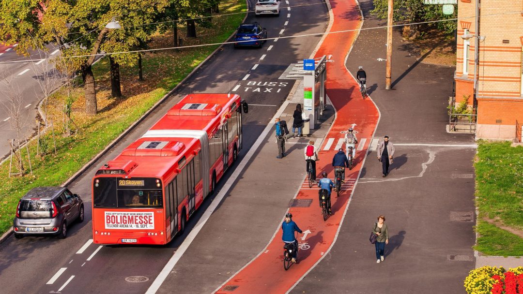 Luftfoto av en rød buss og en taxi på en vei ved siden av et sykkelfelt med syklister og fotgjengere. Trær langs siden, og bygninger er synlige.