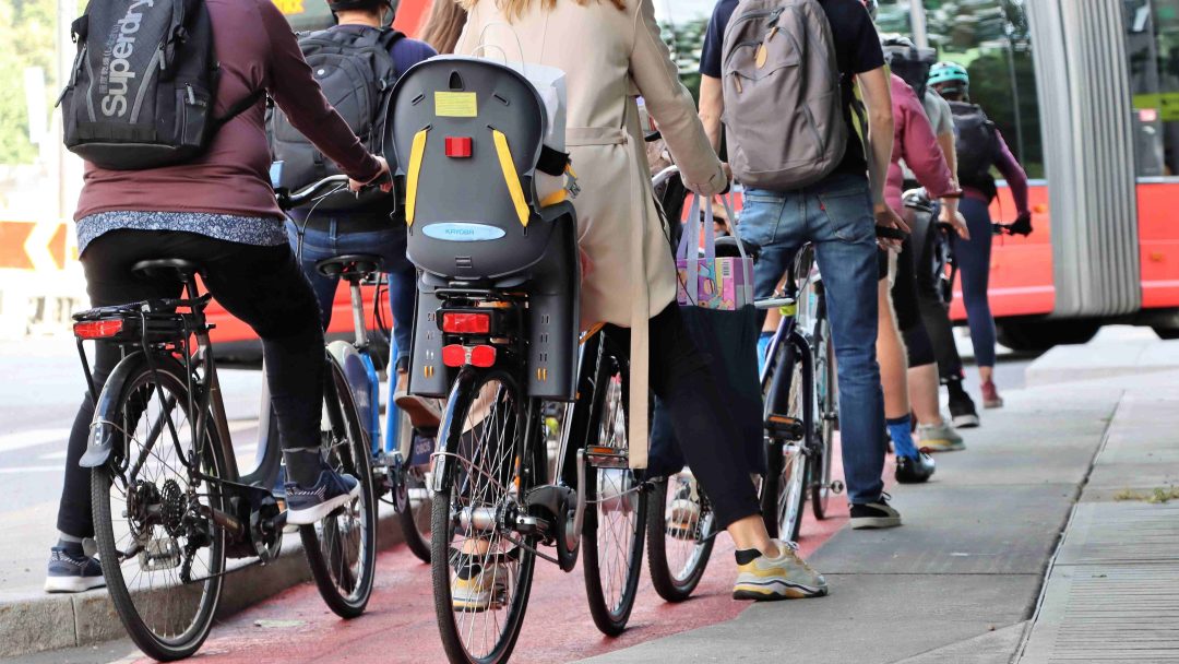 Syklister med ryggsekker og barnevogn på sykkel venter i et anvist kjørefelt bak en rød buss i en bygate.